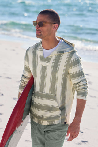 Man on a beach wearing a striped hoodie and sunglasses with a surfboard.