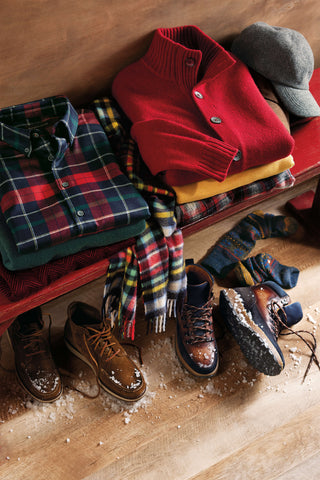Stack of clothing including a red jacket, plaid shirt, and boots on a wooden surface.