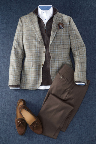 A styled men's outfit laid flat on a blue textured background, featuring a beige and brown plaid blazer over a dark brown zip sweater, a white collared shirt, brown trousers, and brown suede loafers.