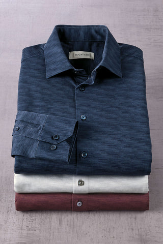 Three neatly folded long-sleeve dress shirts stacked vertically on a textured gray surface. The top shirt is dark navy blue, the middle is off-white/cream, and the bottom is a muted burgundy.