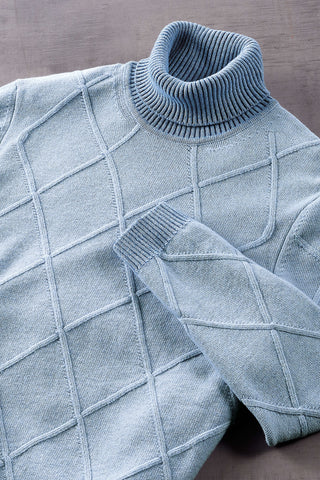 A light blue knit turtleneck sweater with a diamond cable pattern, shown in a close-up flat-lay view on a textured gray surface.