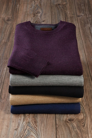 A stack of folded knit crewneck sweaters in multiple colors, with a dark plum purple sweater on top, placed on a rustic wooden surface.