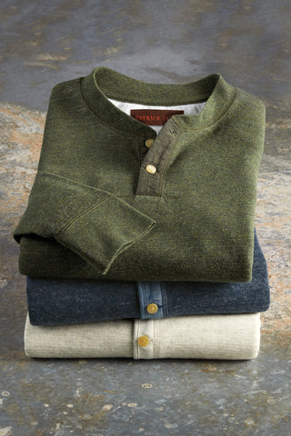 A stack of three folded long-sleeve henley shirts on a textured gray stone surface. The top shirt is olive green, the middle is dark blue, and the bottom is light beige.