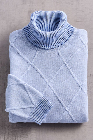 A folded light blue knit turtleneck sweater with a diamond argyle pattern, displayed on a textured gray surface.