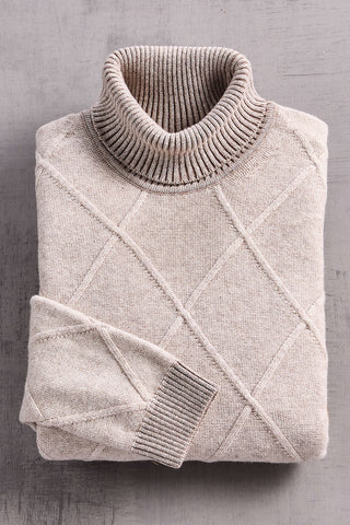 A folded knit turtleneck sweater in a light oatmeal beige color, featuring a textured diamond pattern and ribbed collar and sleeve cuffs, laid flat on a gray textured surface.