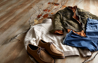 A styled flat-lay of men's fall/winter clothing on a light wood floor, featuring a green waxed cotton jacket, a cream knit sweater, blue jeans, tan suede shoes, and an orange-brown shirt, accented by bare branches with autumn leaves.